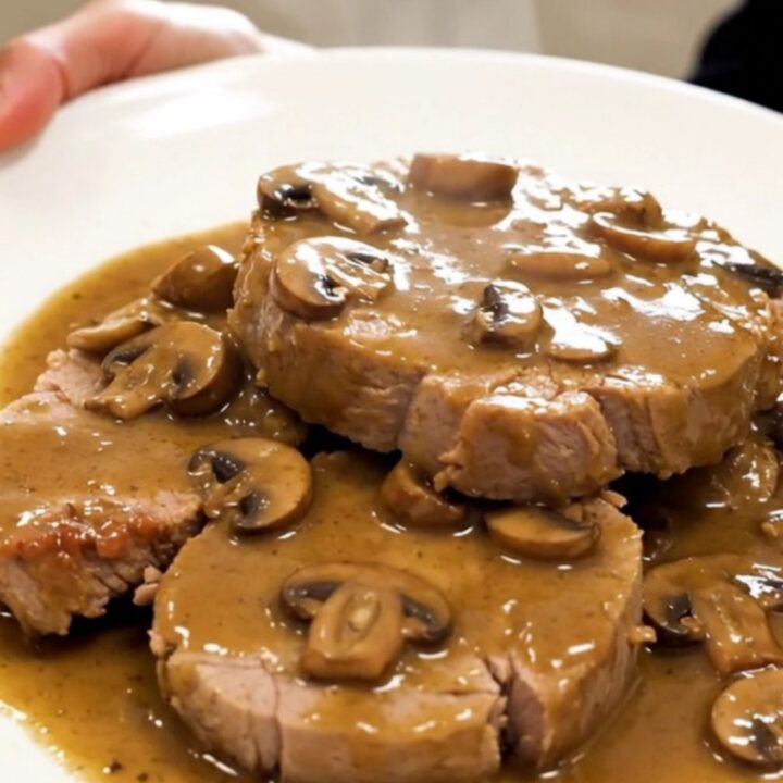 A tender, slow-cooked pork roast topped with a creamy mushroom and shallot sauce on a white plate.
