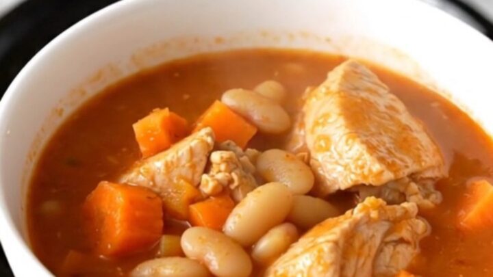 A white bowl filled with steaming chicken and white bean stew on a speckled quartz countertop.