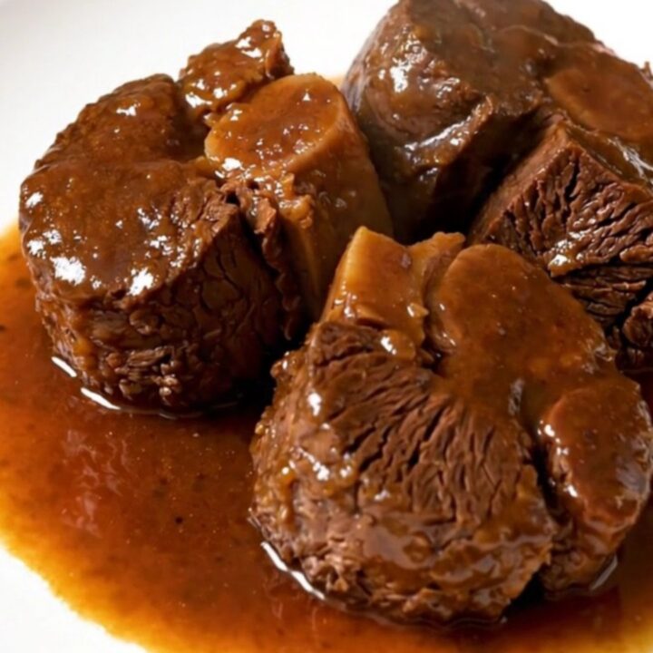 A close-up of tender, braised beef cheeks served on a white plate with carrots and a rich, glossy sauce.