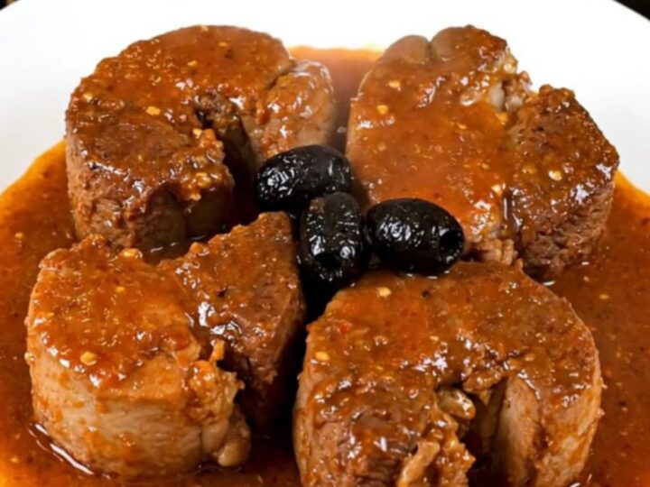 A rustic bowl of tender slow-cooked pork cheeks in a rich tomato sauce with black olives.