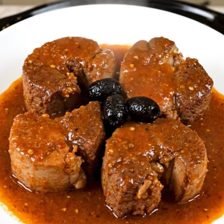 A rustic bowl of tender slow-cooked pork cheeks in a rich tomato sauce with black olives.