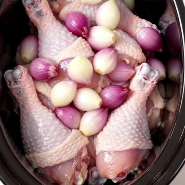 A close-up of tender chicken thighs covered in a glossy red wine glaze with whole shallots on a white plate.