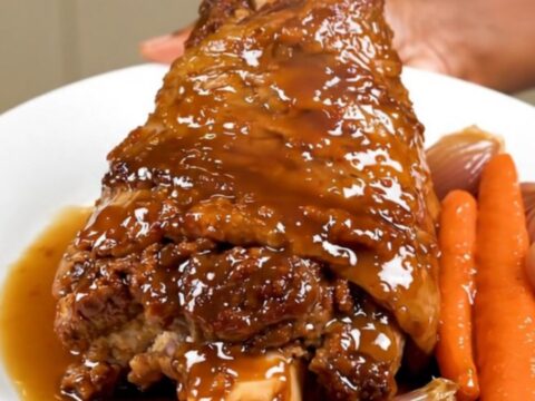 A slow-cooked, tender pork shank served on a white plate with glazed shallots and carrots.