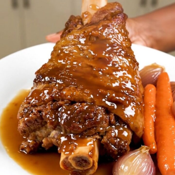 A slow-cooked, tender pork shank served on a white plate with glazed shallots and carrots.