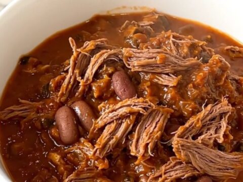 A top-down view of a black slow cooker filled with raw beef chuck roast, sliced onions, kidney beans, and spices.