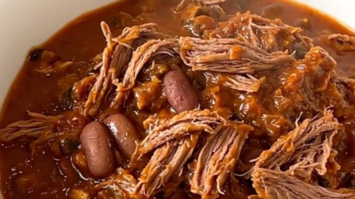 A top-down view of a black slow cooker filled with raw beef chuck roast, sliced onions, kidney beans, and spices.