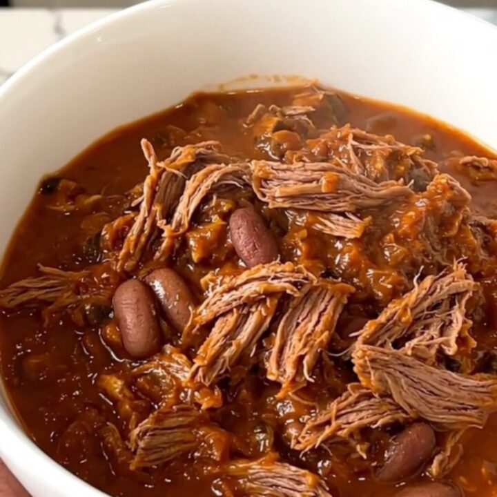 A top-down view of a black slow cooker filled with raw beef chuck roast, sliced onions, kidney beans, and spices.