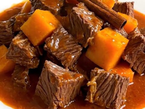 A top-down view of raw beef cubes, diced butternut squash, sliced onions, and a cinnamon stick inside a black slow cooker.