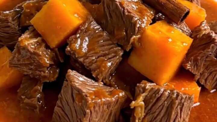 A top-down view of raw beef cubes, diced butternut squash, sliced onions, and a cinnamon stick inside a black slow cooker.
