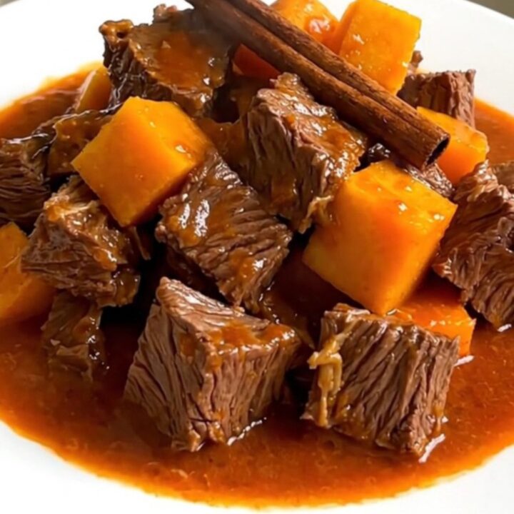 A top-down view of raw beef cubes, diced butternut squash, sliced onions, and a cinnamon stick inside a black slow cooker.