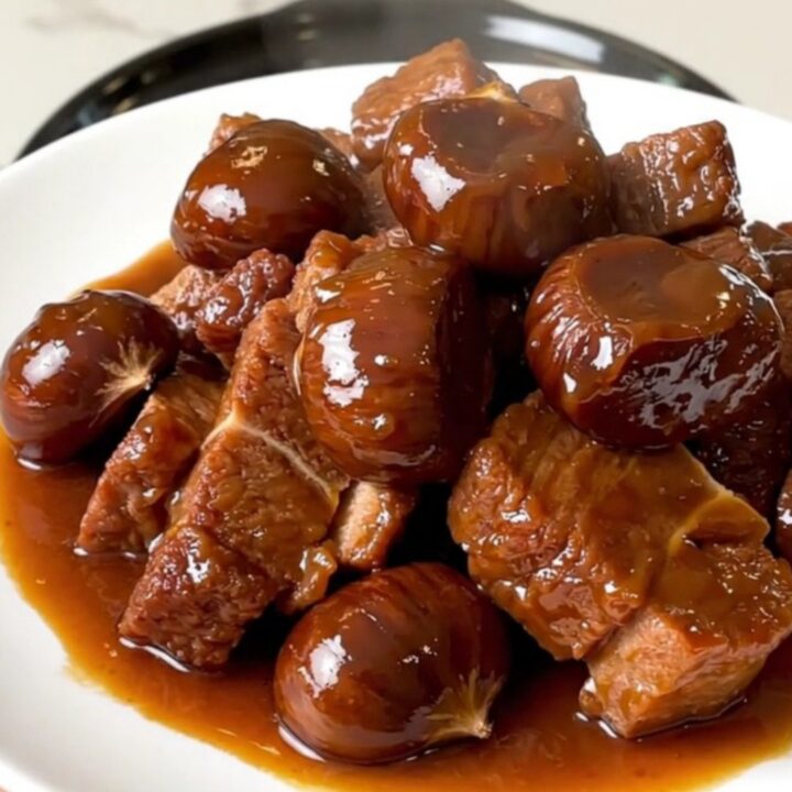A close-up of tender pork collar chunks and whole chestnuts in a rich brown sauce served on a white plate.