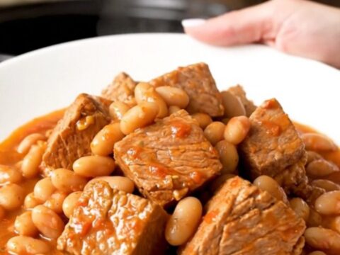 A rustic, steaming bowl of slow-cooked pork shoulder with tender white beans and a rich tomato sauce.