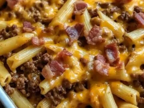 A close-up of a cheesy bacon cheeseburger pasta bake in a ceramic dish, showing a gooey cheese pull with penne.