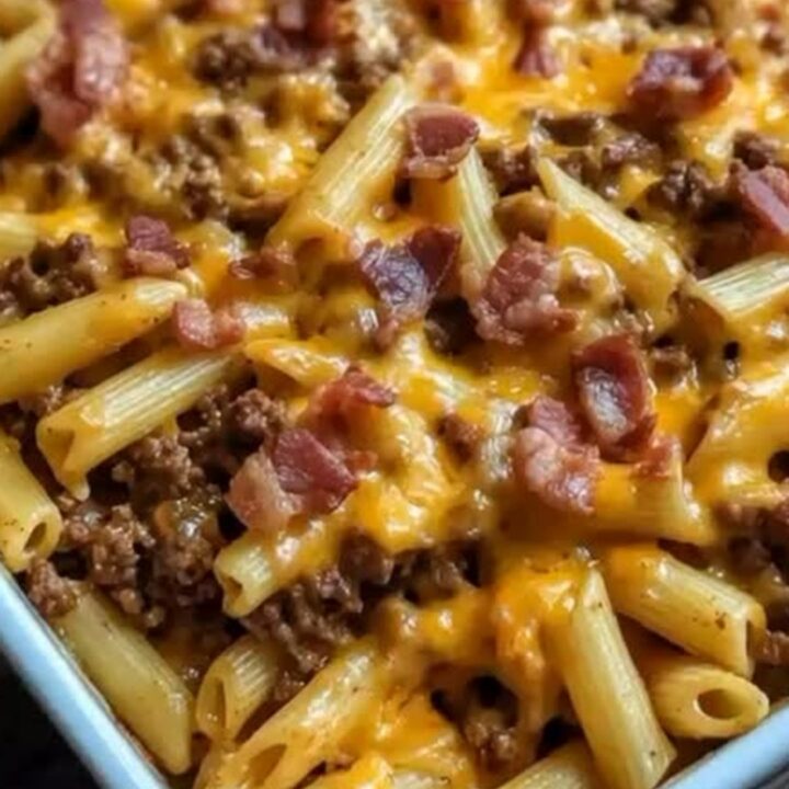A close-up of a cheesy bacon cheeseburger pasta bake in a ceramic dish, showing a gooey cheese pull with penne.