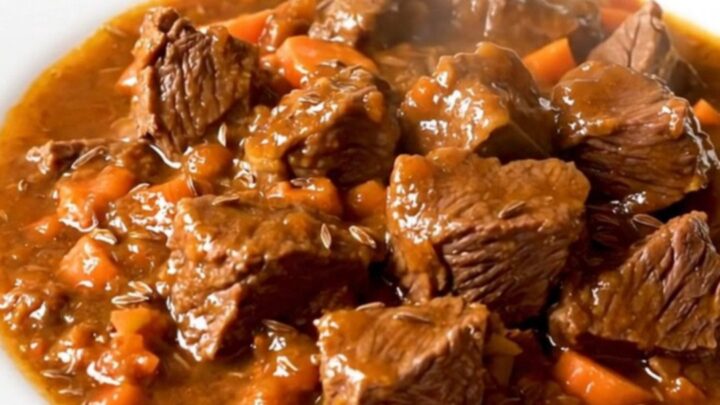 A steaming, glossy beef goulash served on a plain white round plate, showing tender beef chunks and bright orange carrots.