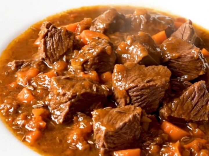 A steaming, glossy beef goulash served on a plain white round plate, showing tender beef chunks and bright orange carrots.