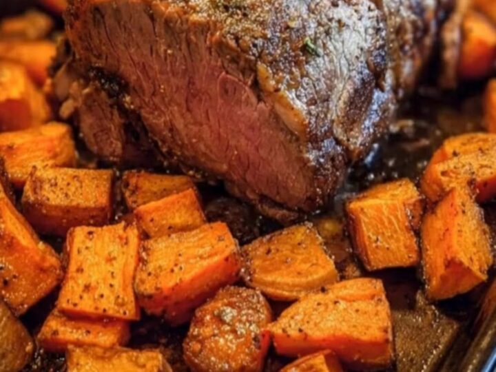 A beautifully sliced herb-crusted roast beef surrounded by golden roasted sweet potatoes on a dark baking pan.