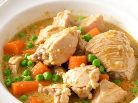 A top-down view of a slow cooker filled with chicken thighs, sliced carrots, peas, and a creamy white sauce.