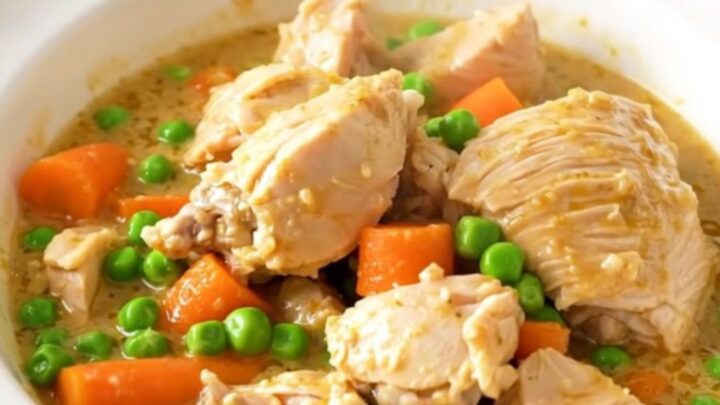 A top-down view of a slow cooker filled with chicken thighs, sliced carrots, peas, and a creamy white sauce.
