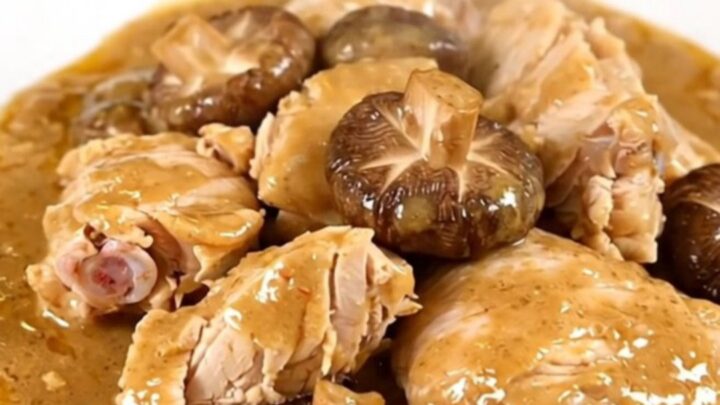 A top-down view of a slow cooker filled with raw chicken thighs, sliced shiitake mushrooms, and broth.