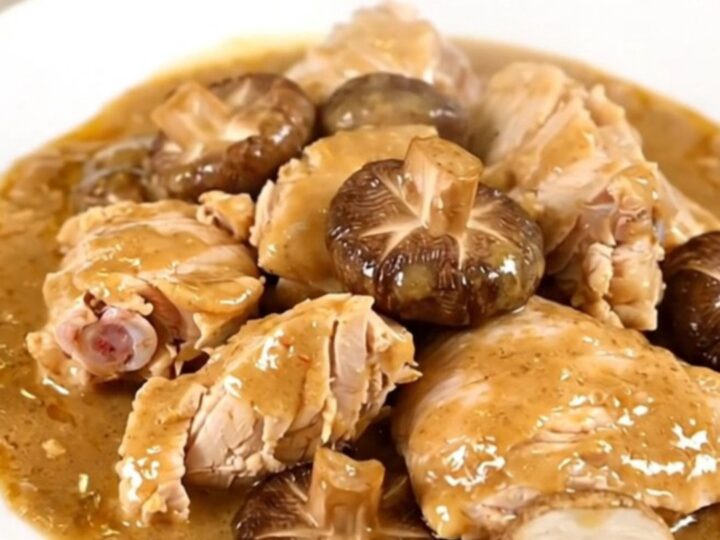 A top-down view of a slow cooker filled with raw chicken thighs, sliced shiitake mushrooms, and broth.