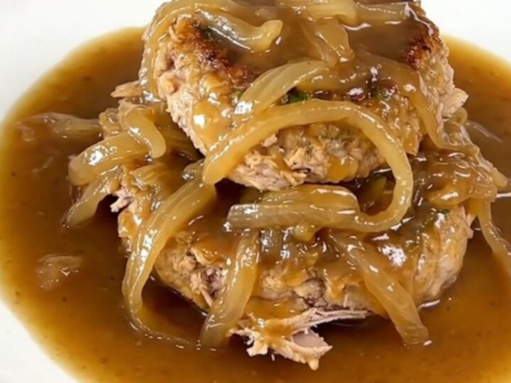 A close-up of slow cooker French onion turkey burger steaks with onions and gravy on a white plate.