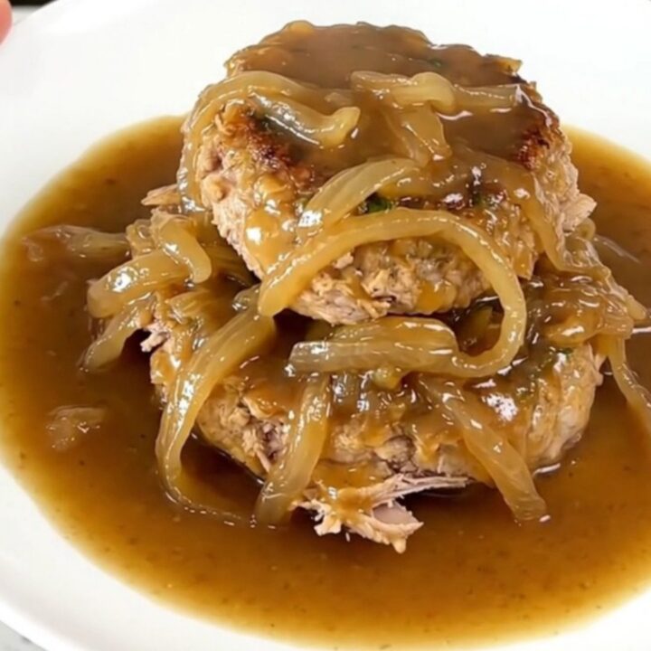 A close-up of slow cooker French onion turkey burger steaks with onions and gravy on a white plate.