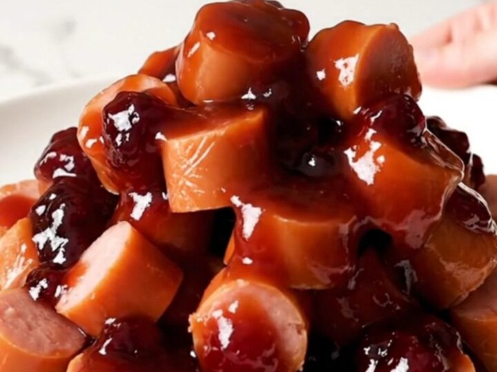 A slow cooker filled with sliced hot dog coins coated in a glossy, dark purple grape jelly barbecue sauce.