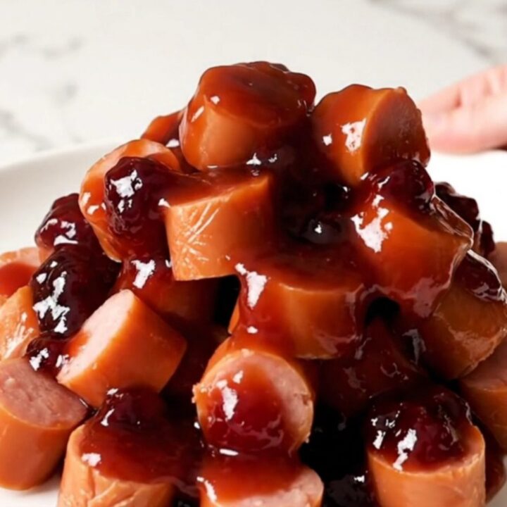 A slow cooker filled with sliced hot dog coins coated in a glossy, dark purple grape jelly barbecue sauce.