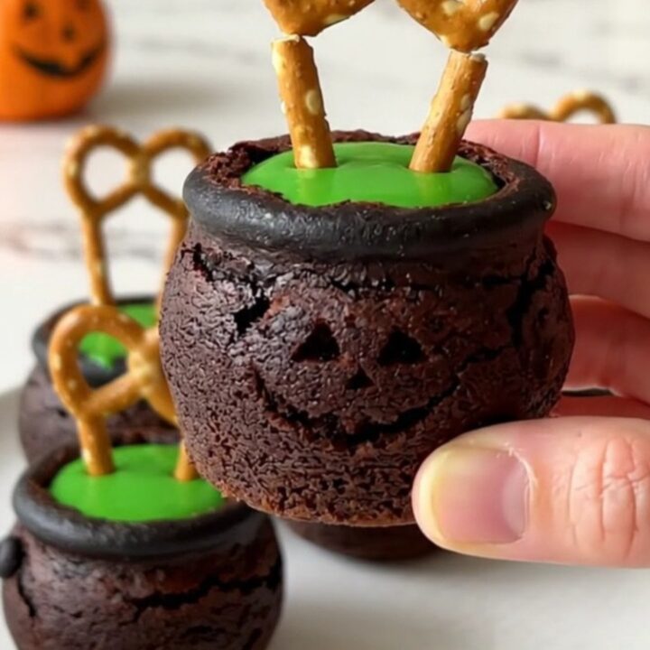A dozen brownie bite cauldrons filled with green candy and pretzel handles on a white plate.