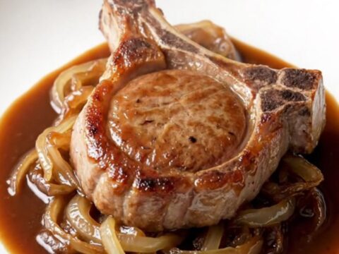 A close-up of a tender, bone-in BBQ pork chop plated on a white plate with a rich, glossy sauce and soft onions.
