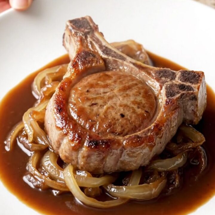 A close-up of a tender, bone-in BBQ pork chop plated on a white plate with a rich, glossy sauce and soft onions.