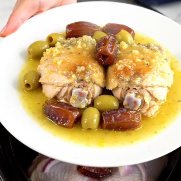 A top-down view of six raw chicken thighs arranged in a black slow cooker insert on a white speckled quartz surface.