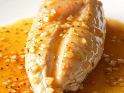 A close-up shot of cooked chicken breasts in a dark slow cooker, coated in a thick, amber honey garlic sauce.