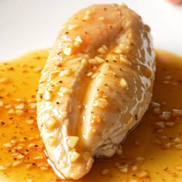 A close-up shot of cooked chicken breasts in a dark slow cooker, coated in a thick, amber honey garlic sauce.