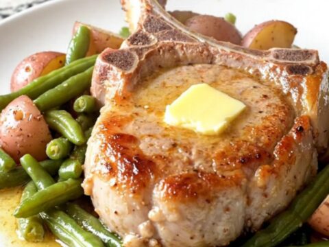 A plate featuring a tender, braised bone-in pork chop with soft green beans and baby potatoes in a light butter sauce.