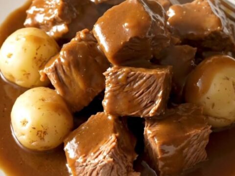 A close-up of tender beef tips and baby potatoes in a rich, savory brown au jus gravy served in a white bowl.