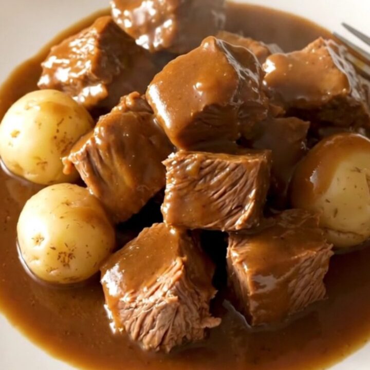A close-up of tender beef tips and baby potatoes in a rich, savory brown au jus gravy served in a white bowl.