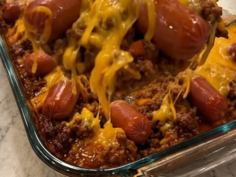 A clear glass baking dish filled with a baked chili cheese dog casserole showing a stretchy cheese pull.