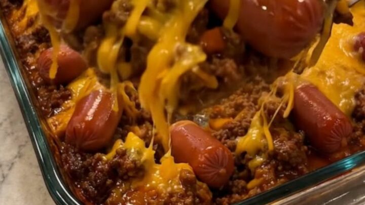 A clear glass baking dish filled with a baked chili cheese dog casserole showing a stretchy cheese pull.