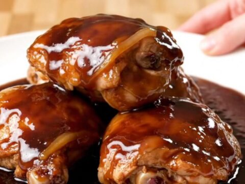 A close-up of slow-cooked chicken thighs in a rich, dark cherry cola glaze on a white plate.