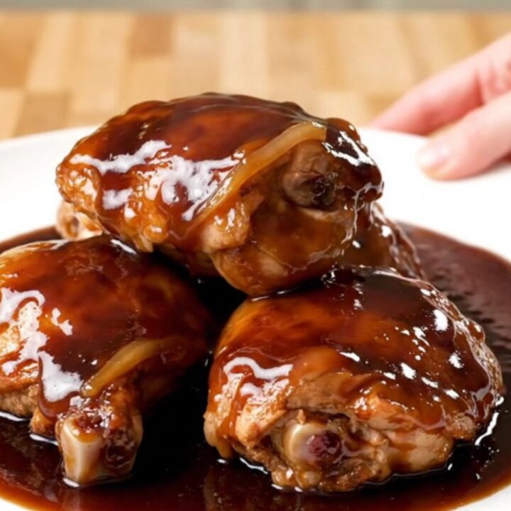 A close-up of slow-cooked chicken thighs in a rich, dark cherry cola glaze on a white plate.