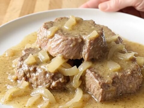 A close-up of slow-cooked cube steaks smothered in creamy, peppered gravy and onions on a white plate.