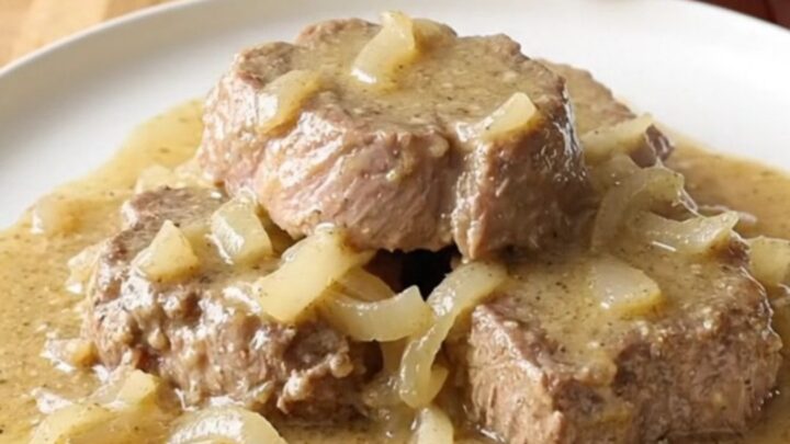 A close-up of slow-cooked cube steaks smothered in creamy, peppered gravy and onions on a white plate.