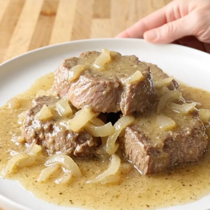 A close-up of slow-cooked cube steaks smothered in creamy, peppered gravy and onions on a white plate.