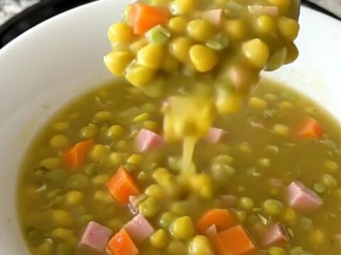 A top-down view of a black slow cooker filled with split pea soup ingredients like ham, carrots, and peas.