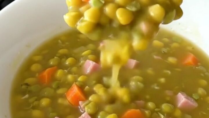 A top-down view of a black slow cooker filled with split pea soup ingredients like ham, carrots, and peas.