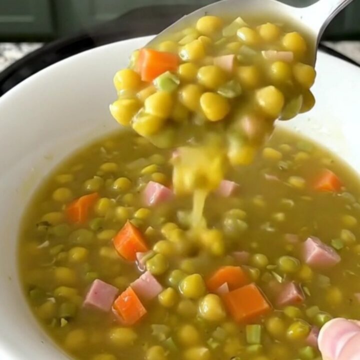 A top-down view of a black slow cooker filled with split pea soup ingredients like ham, carrots, and peas.