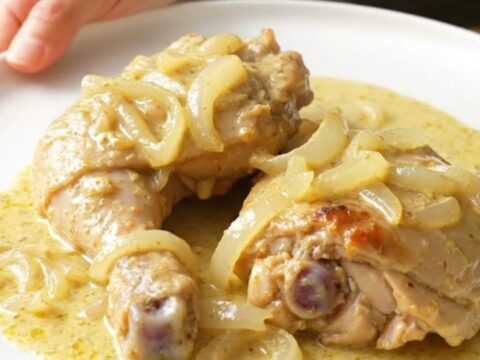 A stainless steel slow cooker filled with tender chicken thighs, soft onions, and a rich, creamy sauce.