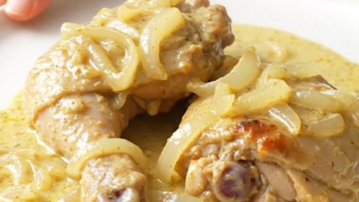 A stainless steel slow cooker filled with tender chicken thighs, soft onions, and a rich, creamy sauce.